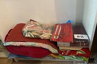 Stack of folded table linens in various colors and patterns, including floral and solid red, rust, and green fabrics.