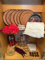 Shelf display showing six rattan charger plates stacked, white and tan napkins, artificial burgundy flowers, dark wood and brass napkin rings, glass bowls, and stainless steel utensils.