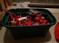 Green plastic bin filled with assorted red and gold Christmas decorations, including baubles and a Santa figurine.