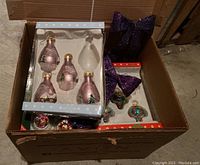 Box filled with boxed pink and clear glass Christmas ornaments and large purple bows