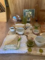 Photo showing variety of vintage pressed glass items including vases, teacups, serving dishes, bowls, and textiles under natural lighting