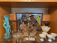 Shelf with various vintage decorative pieces including turquoise deer-shaped vase, clear glass candle holders, white dishes, plated silver candle holders, and framed Vogue mirrored art