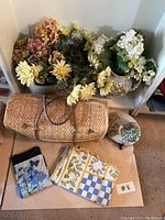 Photo of vintage straw picnic basket, picnic cloths, and multiple artificial flower bunches arranged on a shelf and floor.
