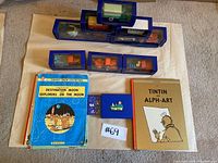 Seven tin toy cars in blue display cases arranged on a surface along with three Tintin books including 'Destination Moon' and 'Tintin Alph-Art'.