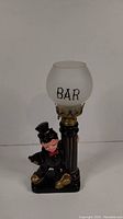 Front view of the 1950's Hobo Bar ceramic oil lamp featuring a hobo figure, black column base with gold trim, and frosted glass globe labeled 'BAR'.