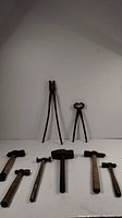 Photo showing two pairs of blacksmith tongs: one large pair with concave jaws and one medium pair with round jaws, along with five hammers and a metal anvil block laid out on surface.