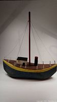 Full view of folk art wooden fishing boat model with blue hull, yellow trim, brown deck, single red mast and small cabin