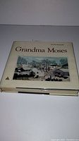 The front cover of the Grandma Moses coffee table book by Otto Kallir, showing a winter landscape illustration on a beige dust jacket.