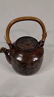 Front angled view of a dark brown ceramic teapot with bamboo handle showing textured surface and lid on top.