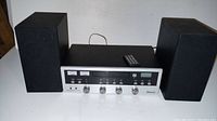 Full front view of stereo system with two speakers and remote on top, showing control panel, knobs, Bluetooth logo.