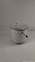Front and side angle of the white enamel pot with red rim and wire handle, showing minor lip chips.
