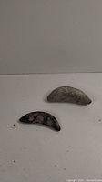 Two crescent-shaped stone axe heads on a white surface, one larger gray with speckles, one smaller dark with patches.