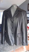 Front view of black leather ladies jacket on a dress form, showing button front closure, flap pockets, and collar.