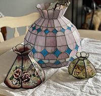 Three stained glass shades displayed on a table, including the large pink and blue hanging light and two smaller painted glass shades