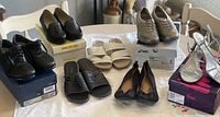 Overall view of 7 pairs of shoes arranged on table showing various styles including leather dress shoes, slides, sneakers, flats, and glittery heels