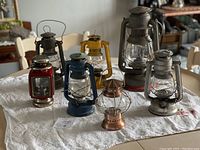 Seven vintage lanterns of different colors and designs arranged on a cloth sheet, including oil, candle, and battery-powered types