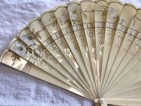 Close view of the hand fan showing painted floral design, perforated celluloid sticks, and scalloped edges.