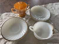 Photo showing four white milk glass medium-sized plates, one white milk glass handled sugar bowl with spout, and one amber glass two-handled sugar bowl with embossed decorative pattern, placed on cloth surface.