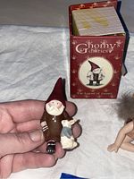 Photo showing the Gnomy's Diaries gnome figurine held in hand with its original box in the background and a small porcelain fairy figurine nearby.