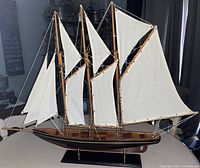 Full view of the wooden sailboat model showing multiple cream-colored fabric sails and rigging with black hull on display stand.