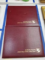 Two burgundy leatherette albums stacked, showing gold embossed Reader's Digest First Day Cover Collection text and logo on covers.