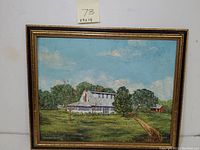 Framed oil painting showing homestead scene with white farmhouse, trees, and dirt path under blue sky.