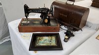 Antique Singer sewing machine on wooden base, vintage embroidery art, and brown leather-like locking case with power cord and foot pedal on table.