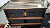Front and top views of the antique steamer trunk showing metal bands, wooden slats, caster wheels, and padlock closure.