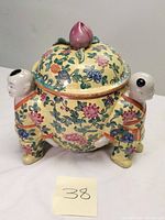 Front and side view of the jar showing the yellow floral body and attached child figures forming legs, with a pink peach finial on the lid.