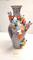 Front view showing five colorful children figurines climbing and holding an orange-brown tree trunk on a blue and white floral patterned vase with a brown rim.