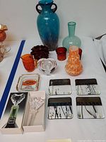 Wide view of the collection displaying multiple art glass vases, a clear glass paperweight, textured red and amber glass pieces, and glass tile coasters.