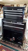 Two stacked black Craftsman tool cabinets, top chest and bottom rolling cabinet on a carpeted floor.