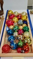 Top-down view of approximately 26 assorted vintage glass Christmas ball ornaments arranged in a wooden tray show a variety of colors including shiny gold, red, pink, silver, blue, green, cream, and orange. Several feature reflective metallic finishes and disco ball styles.
