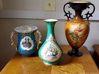 Three vintage European porcelain vases displayed on a wooden surface, showing front views, artistic details and colors.