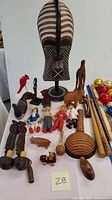 Front view of a large carved tribal wooden mask on stand with various smaller carved wooden figurines and souvenirs arranged around it including dolls, animals, and baseball bats.