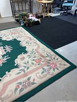 Partial view of large floral rug and black floor mat on basement floor with some shelves and tables in background