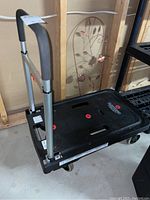 Black plastic flatbed dolly with aluminum extendable handle and four caster wheels shown from an angled side view.