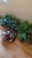 Three different faux plant selections placed on wood floor, including one floral arrangement and two green leafy plants.