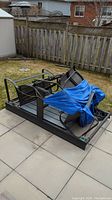Disassembled swing components including metal frame base and chairs covered by tarp on patio