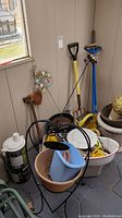 Photo showing garden tools including a yellow shovel, blue hose sprayer, several garden pots, gloves, a decorative metal plant stand with 'Welcome' sign, and a blue watering can.