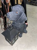 Small black resin dog statue holding a lantern in its mouth on concrete floor, partial view of metal garden decor with leaf accents behind