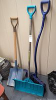 Photo showing three shovels: metal shovel with wooden handle and two plastic snow shovels (blue and purple) with different handles and scoops.