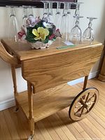 Front side of solid oak tea wagon showing drop leaves and wheels, with glasses and floral arrangement on top