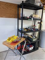 Foldable wooden worktable with handled hand tools and yellow plastic miter box on top, black shelving unit in background with other items.