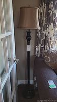 Full view of the 60-inch black metal floor lamp with beige cloth shade beside a couch and door.