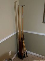 Photo showing six pool cues and cue rake standing upright in a black circular stand against a beige wall.