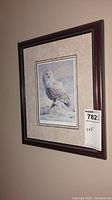 Framed print showing snow owl perched on snow in winter landscape with mountains in the background, hung on wall with lot sticker attached on lower right of frame.