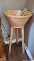 Wooden salad bowl mounted on a three-legged stand, placed near a corner with visible power outlet.