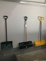 Limited view of three snow shovels standing upright showing overall shapes, handle types and colors