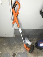 Full view of cordless orange and silver weed trimmer showing handle and body, resting against a door on concrete floor.